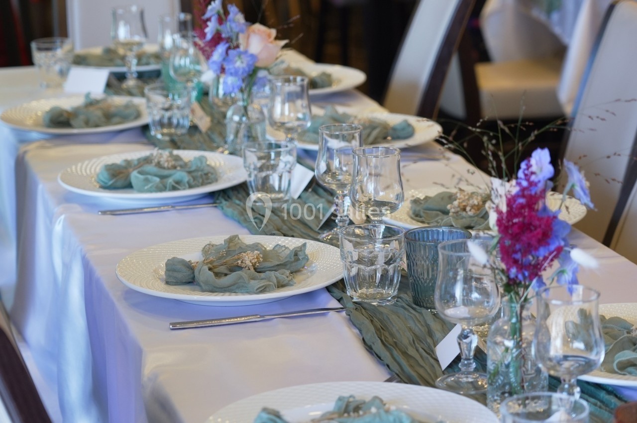 Table dressée avec nappes blanches, serviettes bleues, verres, couverts et décorations florales colorées.