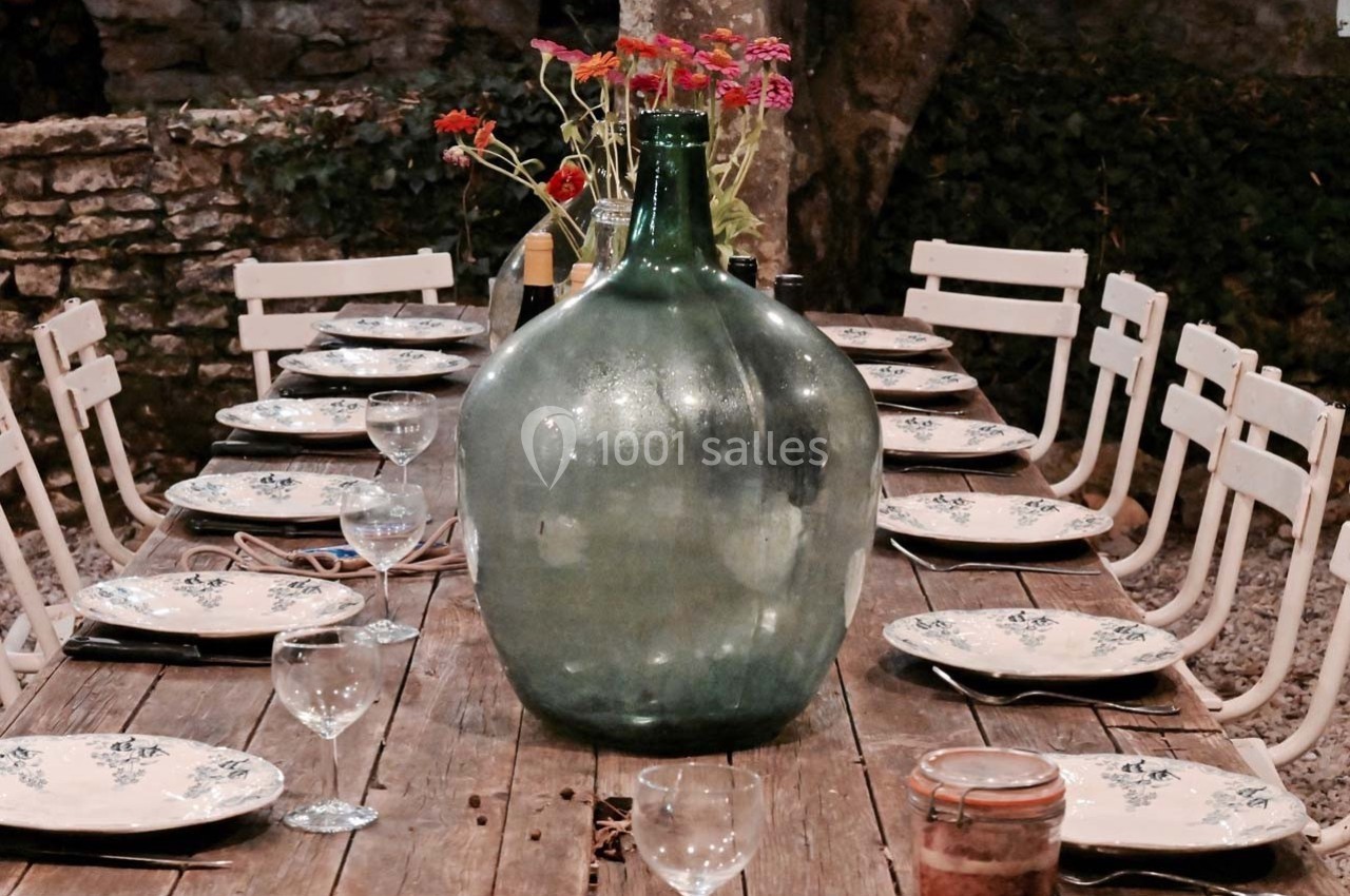 Grande table en bois dressée avec assiettes et verres, décorée d'une bonbonne en verre et de fleurs colorées.