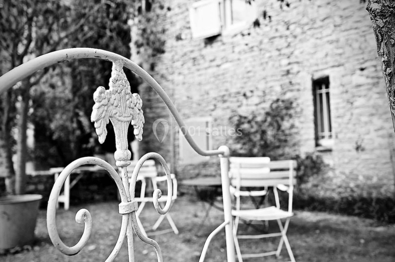 Vue en noir et blanc d'un jardin avec une clôture en fer forgé au premier plan et des chaises devant un mur en pierre.