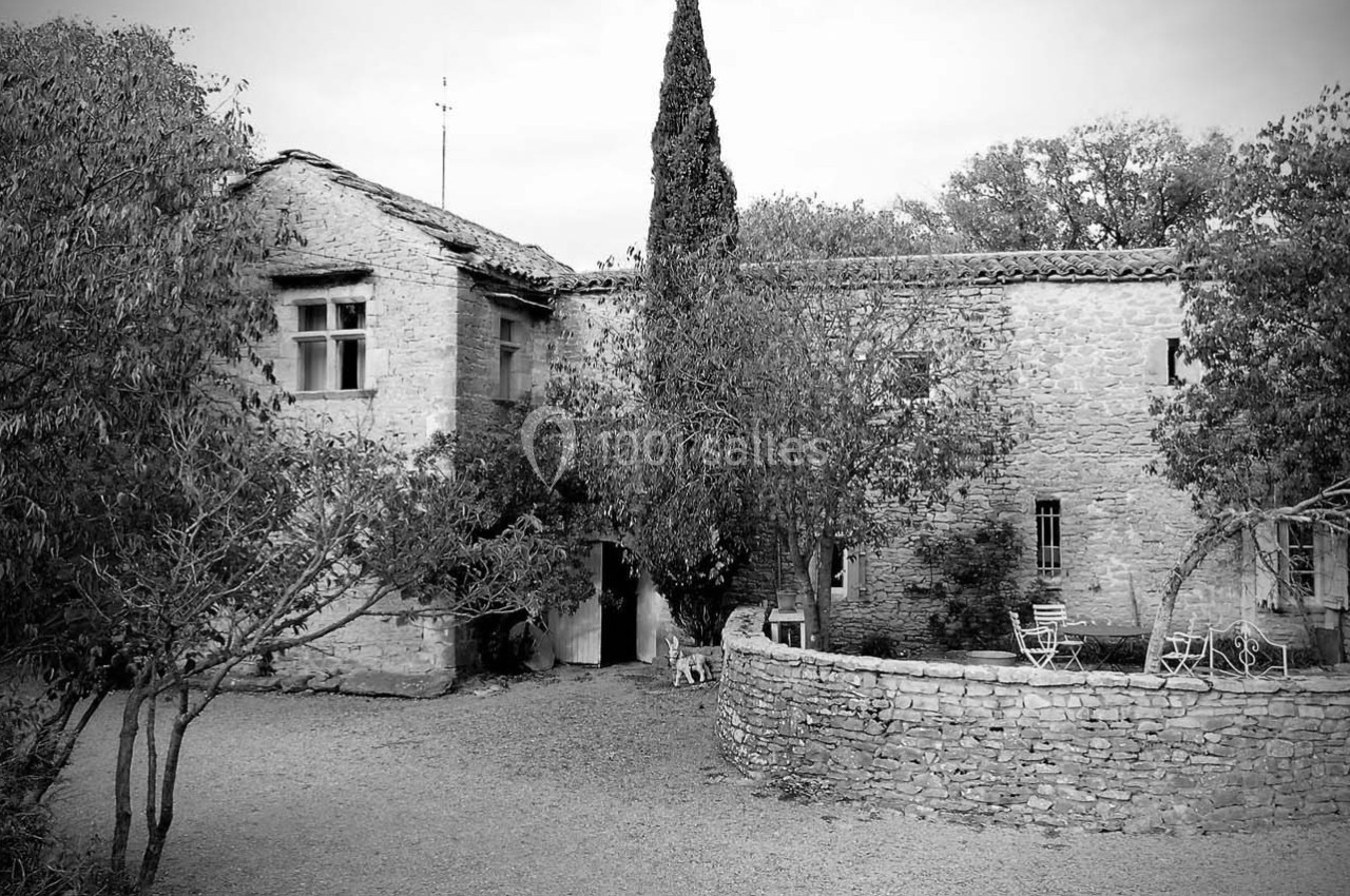 Bâtisse en pierre ancienne entourée d'arbres, avec une cour et un muret en pierre au premier plan.