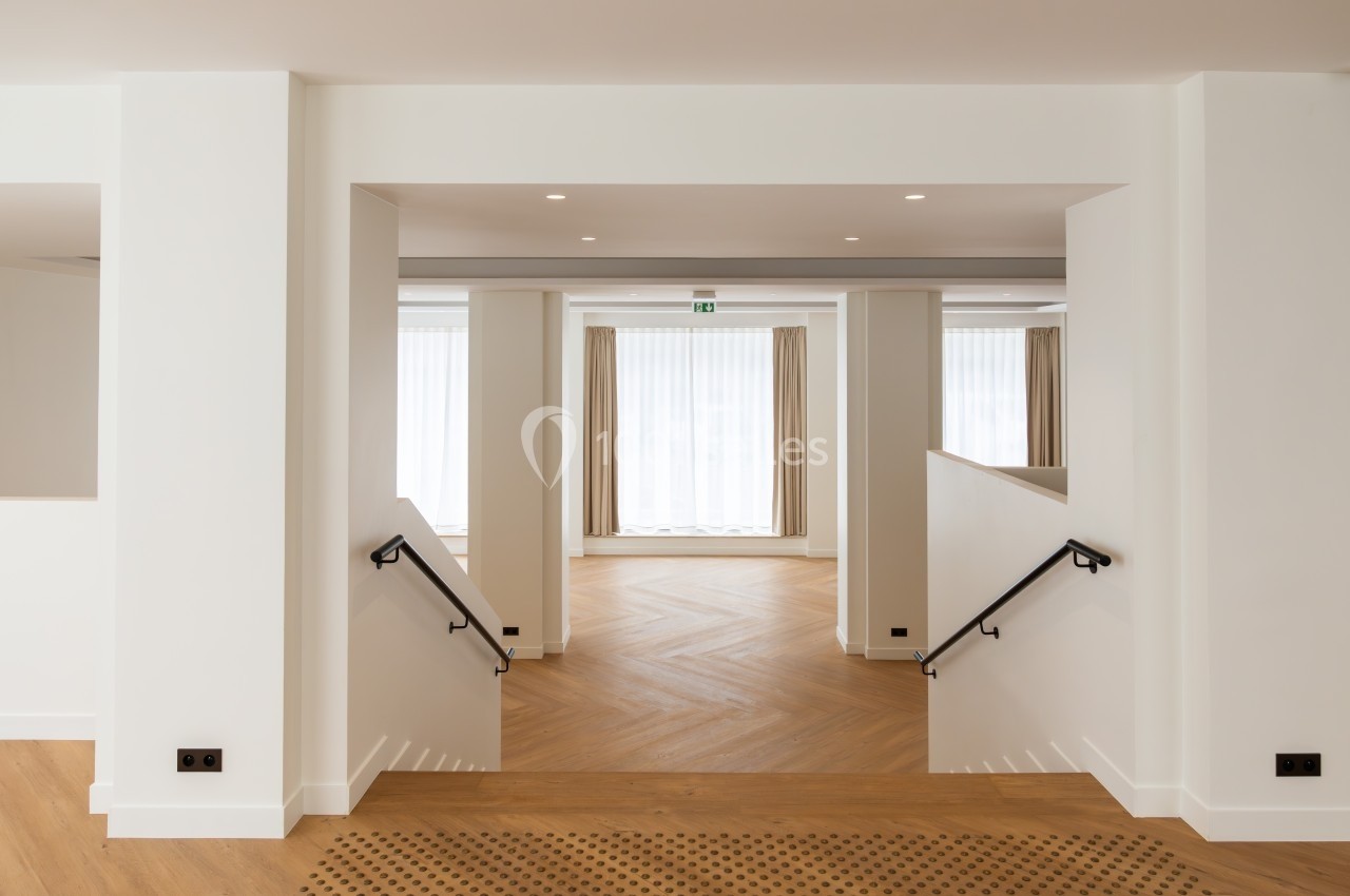 Vue d'un intérieur lumineux avec parquet en chevrons, murs blancs et escalier descendant vers une grande baie vitrée.
