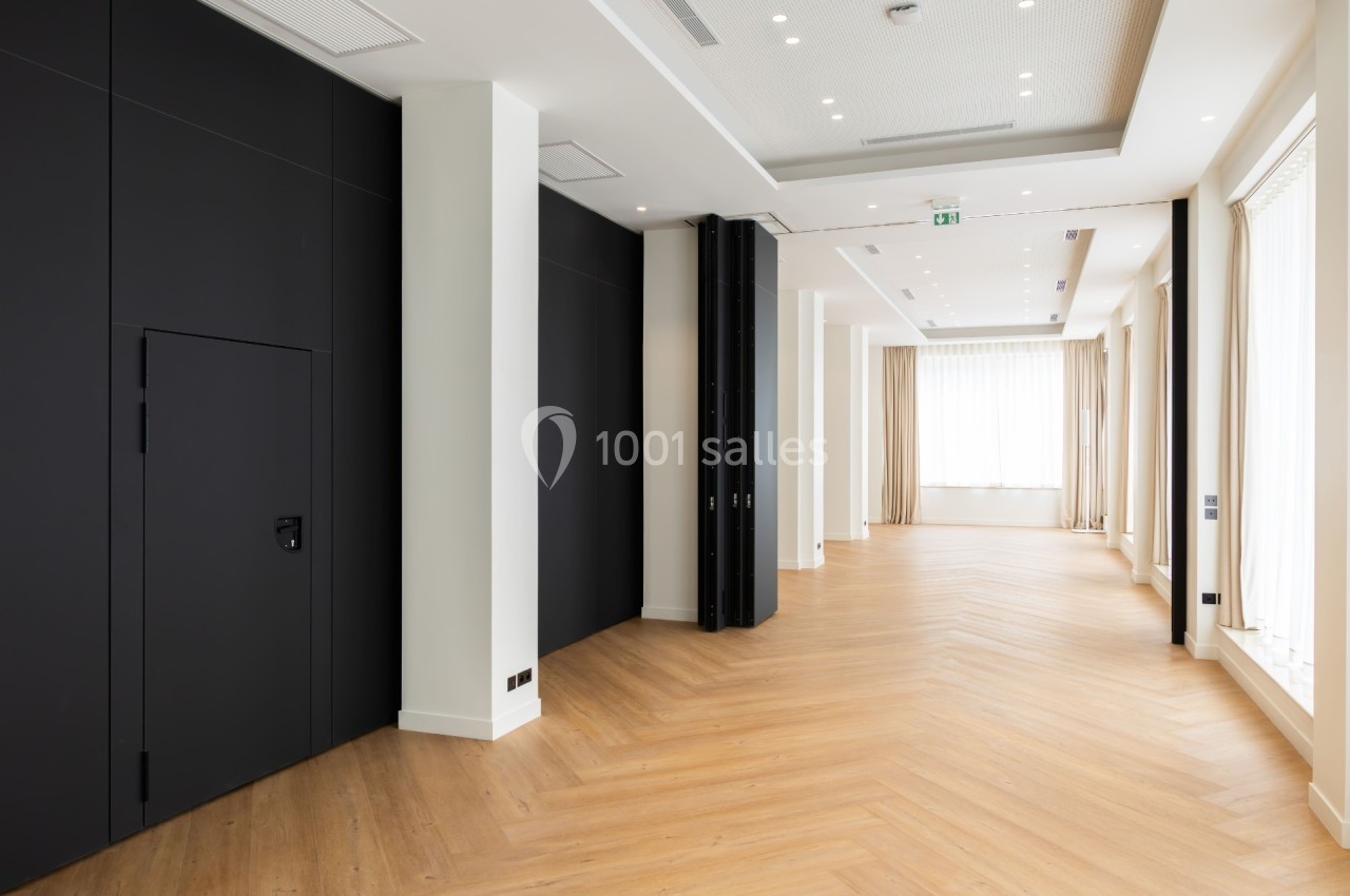 Salle lumineuse avec parquet en chevrons, murs blancs, panneaux noirs et grandes fenêtres laissant entrer la lumière…