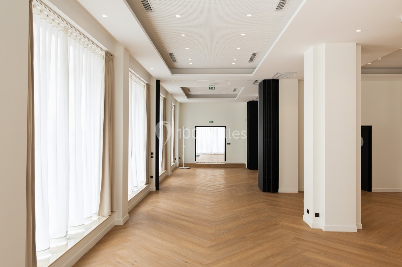 Salle lumineuse avec parquet en bois clair, grandes fenêtres avec rideaux blancs et murs blancs épurés.