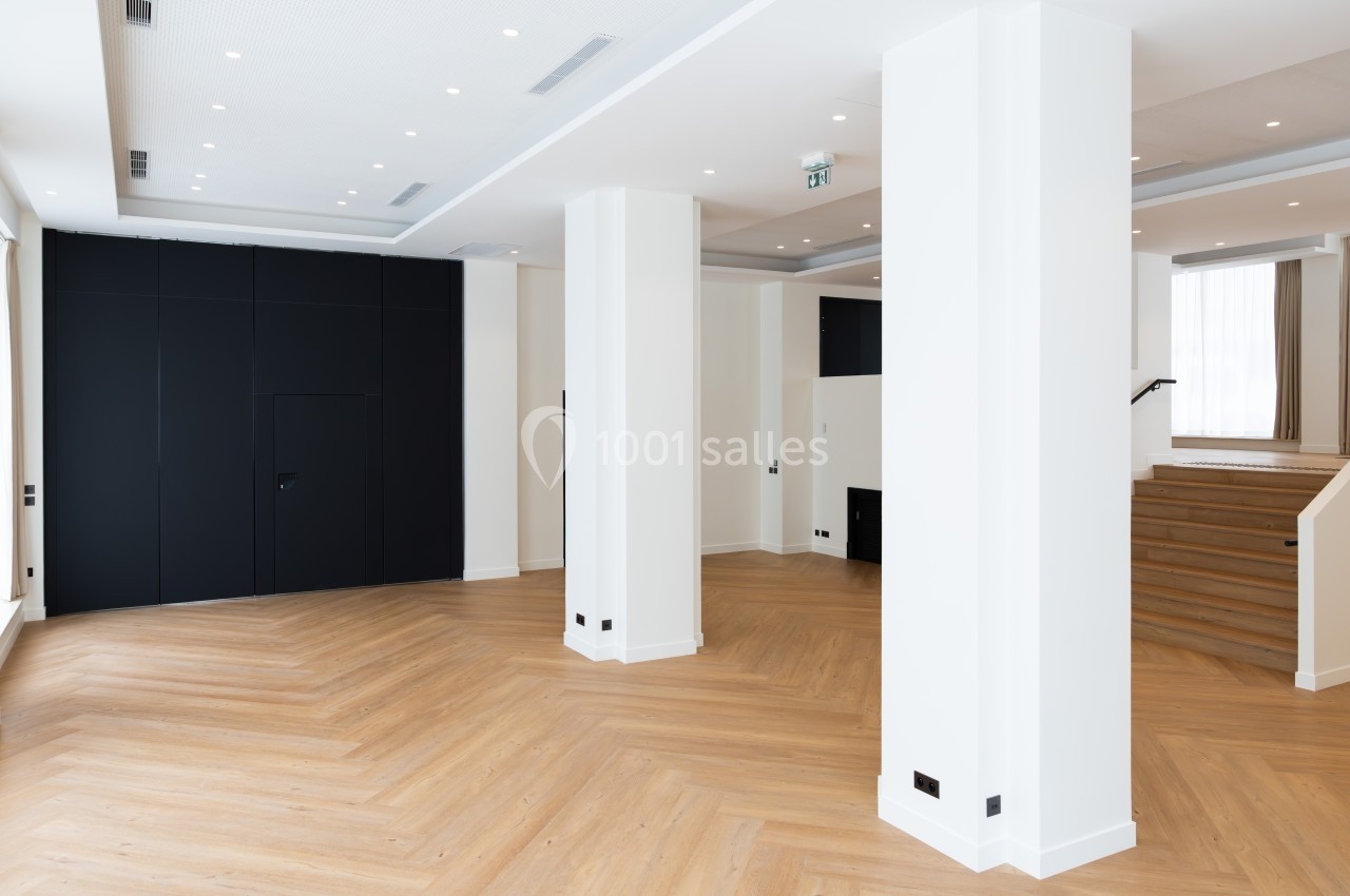 Salle vide avec parquet en bois clair, murs blancs, panneaux noirs et escalier en bois dans un espace lumineux.