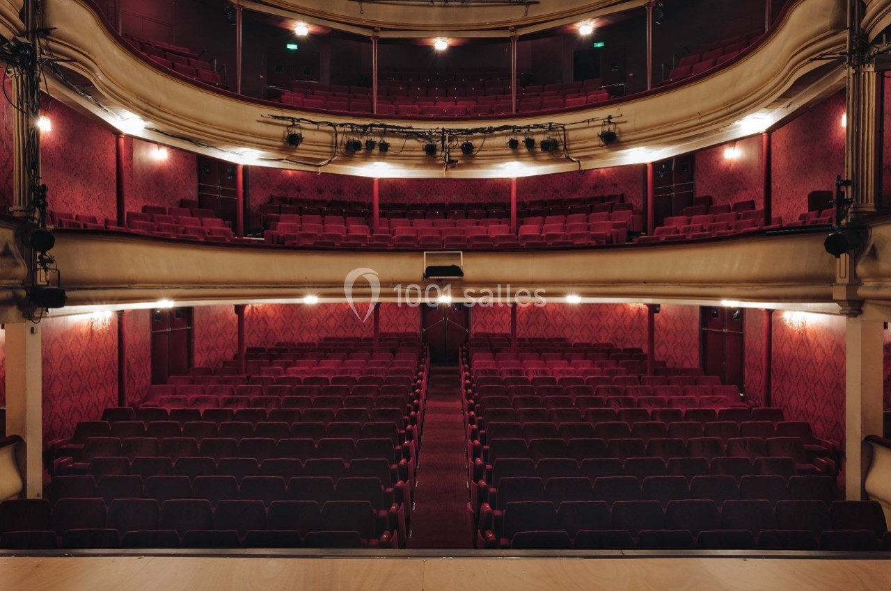 Salle de théâtre vide avec rangées de sièges rouges, balcons ornés et éclairage tamisé.