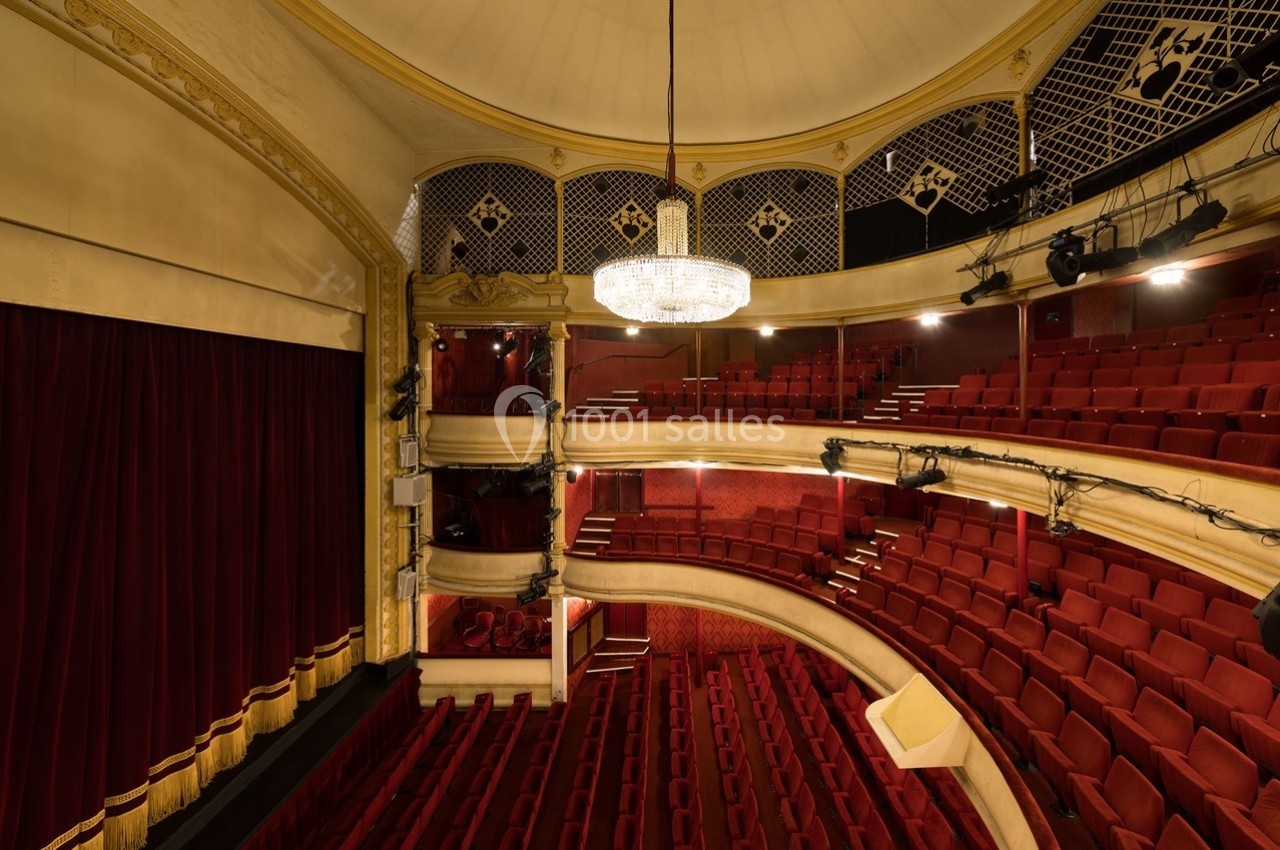Salle de théâtre avec sièges rouges, balcon, rideaux fermés et grand lustre central sous un plafond voûté.