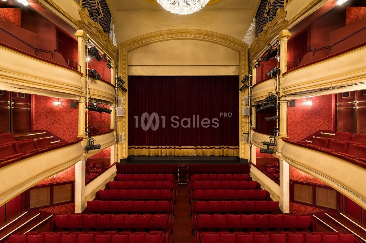 Salle de théâtre avec sièges rouges, balcons ornés et rideau de scène fermé sous un grand lustre.