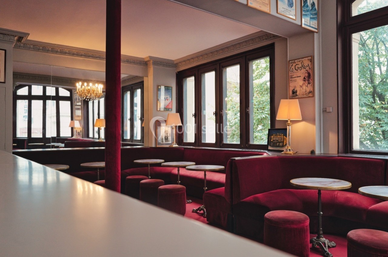 Salle de café élégante avec banquettes rouges, tables rondes en marbre, grandes fenêtres et lustre central.