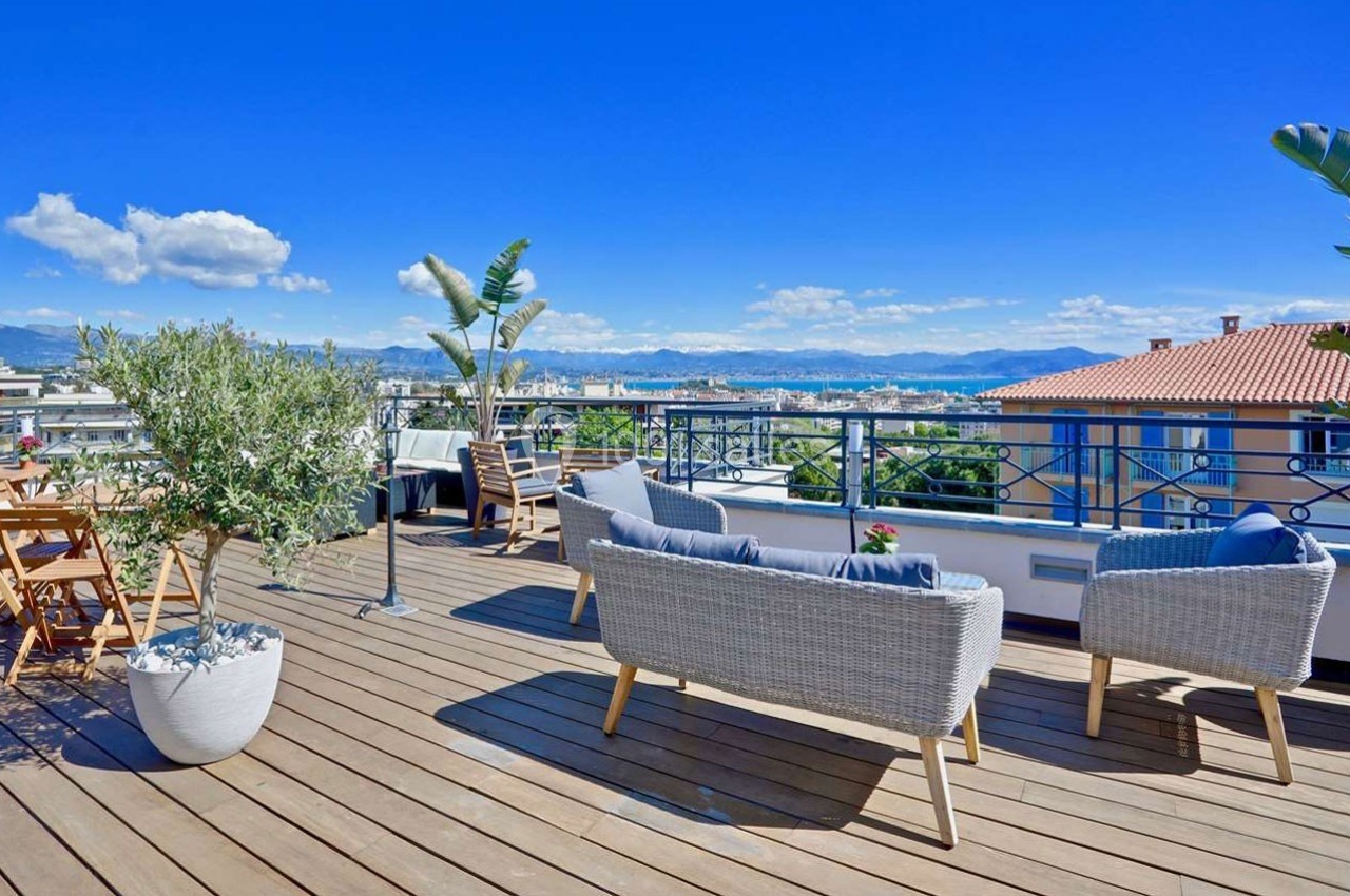 Terrasse en bois aménagée avec mobilier d'extérieur, plantes en pot et vue dégagée sur des toits et des montagnes.