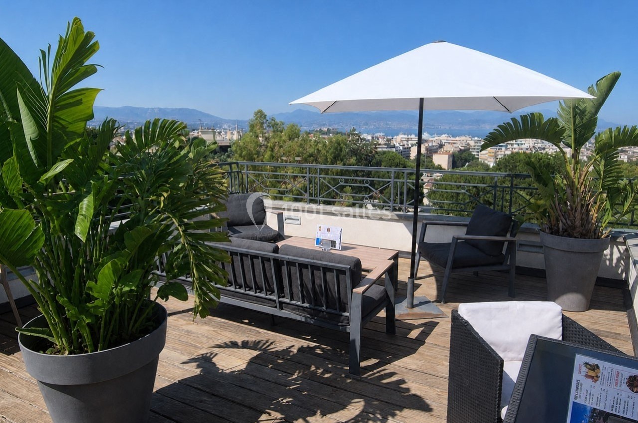 Terrasse ensoleillée avec mobilier de jardin, parasol blanc et plantes en pot, offrant une vue sur une ville et des collines.