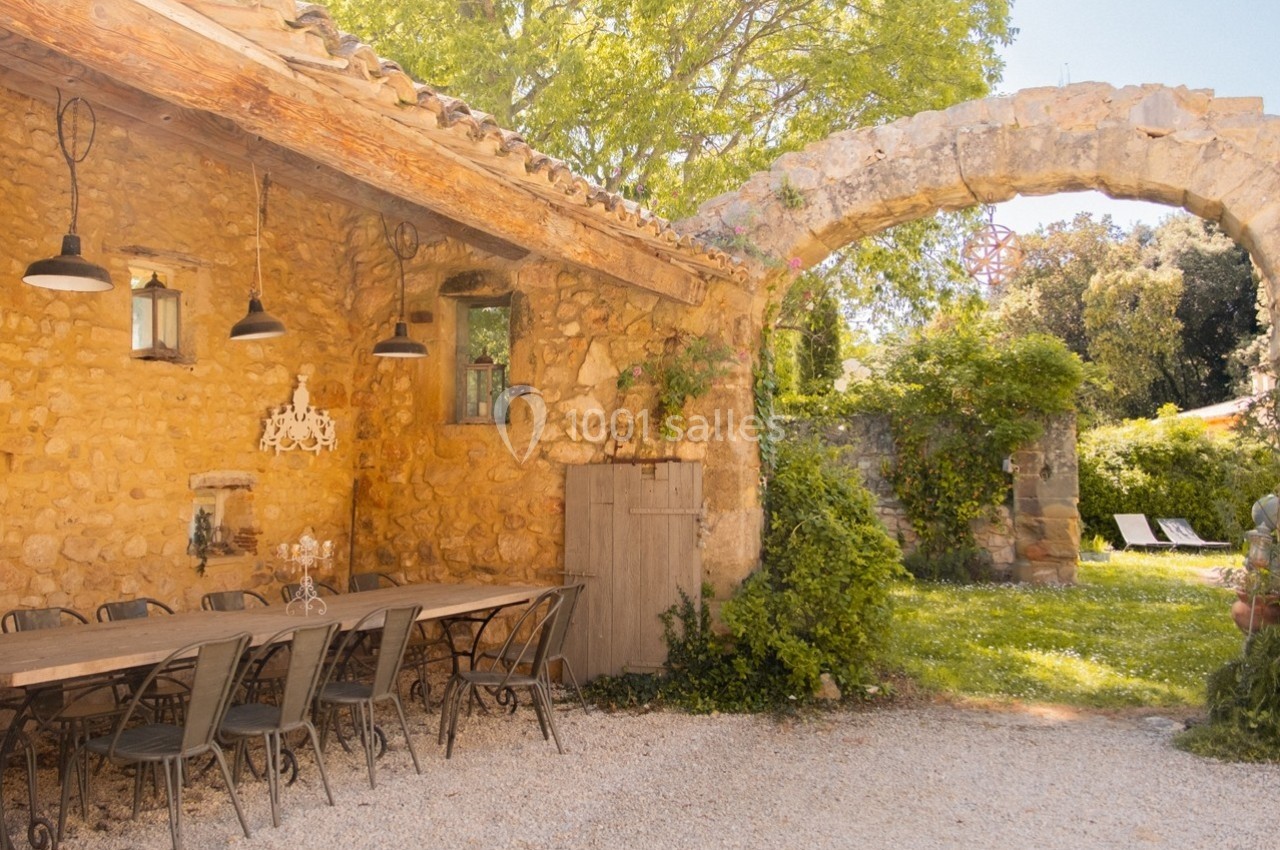 Terrasse rustique avec table en bois, chaises métalliques et arche en pierre dans un jardin verdoyant.