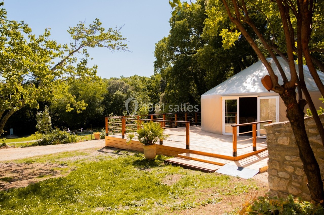Terrasse en bois devant une yourte blanche entourée d'arbres et de verdure sous un ciel dégagé.