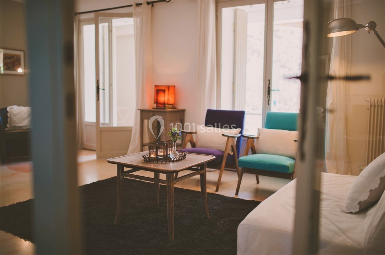 Salon lumineux avec mobilier vintage, tapis noir, table basse en bois et fauteuils colorés près de grandes fenêtres.