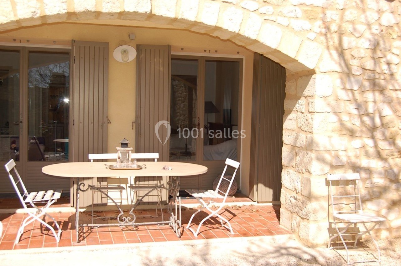 Terrasse en pierre avec table ovale, chaises blanches pliantes et lanterne, devant une maison aux volets fermés.