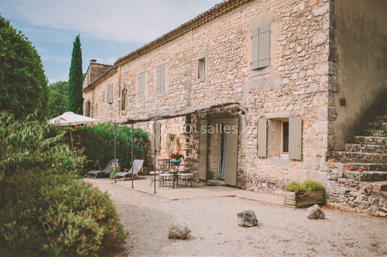 Façade en pierre d'une maison traditionnelle avec terrasse aménagée, parasol et chaises dans un jardin arboré.