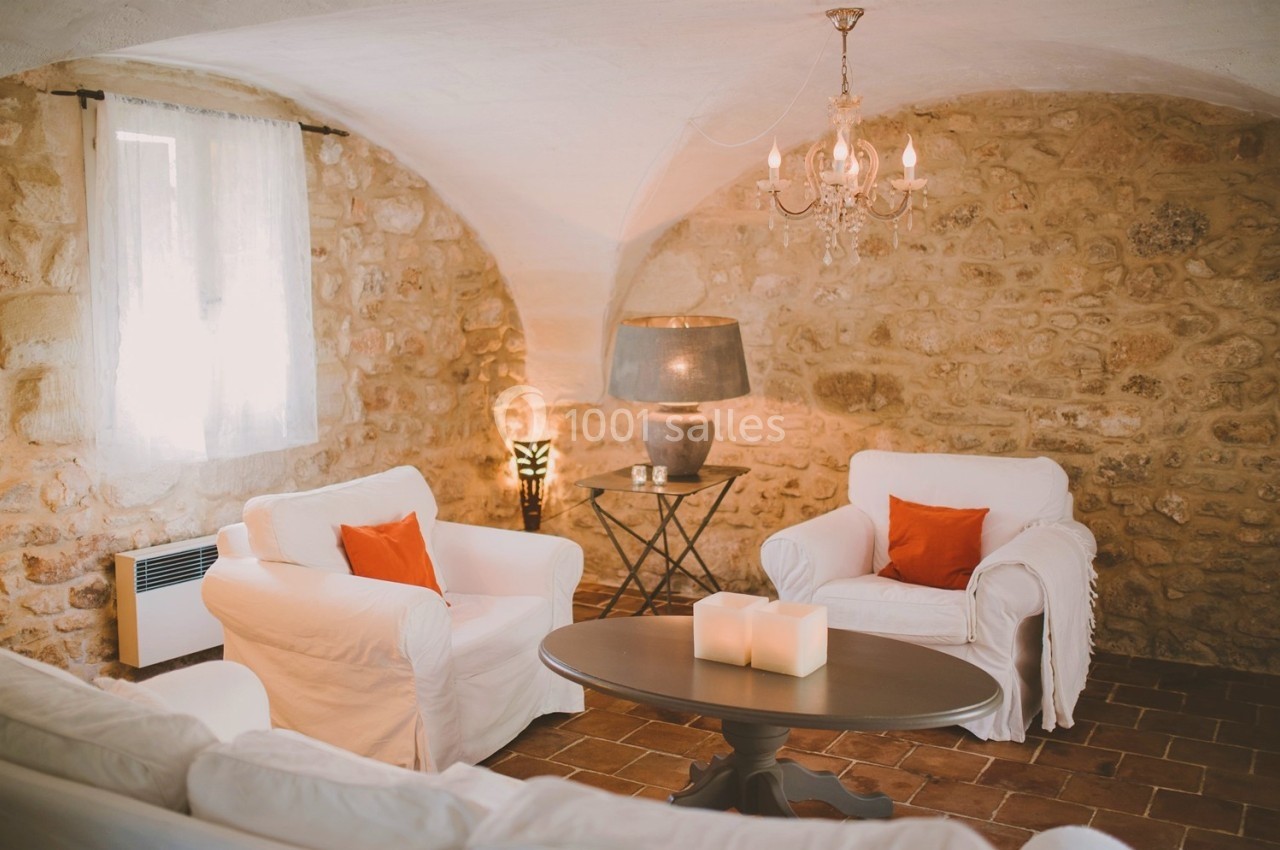 Salon chaleureux avec murs en pierre, fauteuils blancs, coussins orange, table basse et éclairage tamisé.