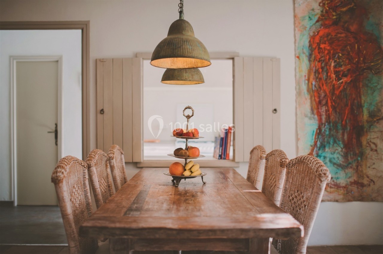Table en bois avec des chaises en osier, plateau de fruits au centre, éclairée par une suspension dans une pièce décorée.