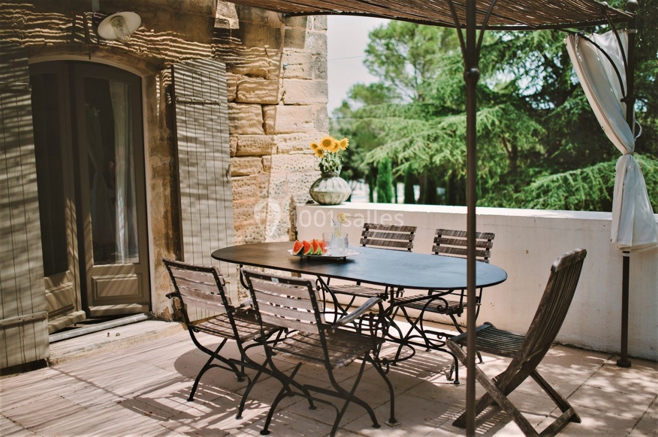 Terrasse ensoleillée avec table et chaises en métal, vase de tournesols et vue sur des arbres verdoyants.