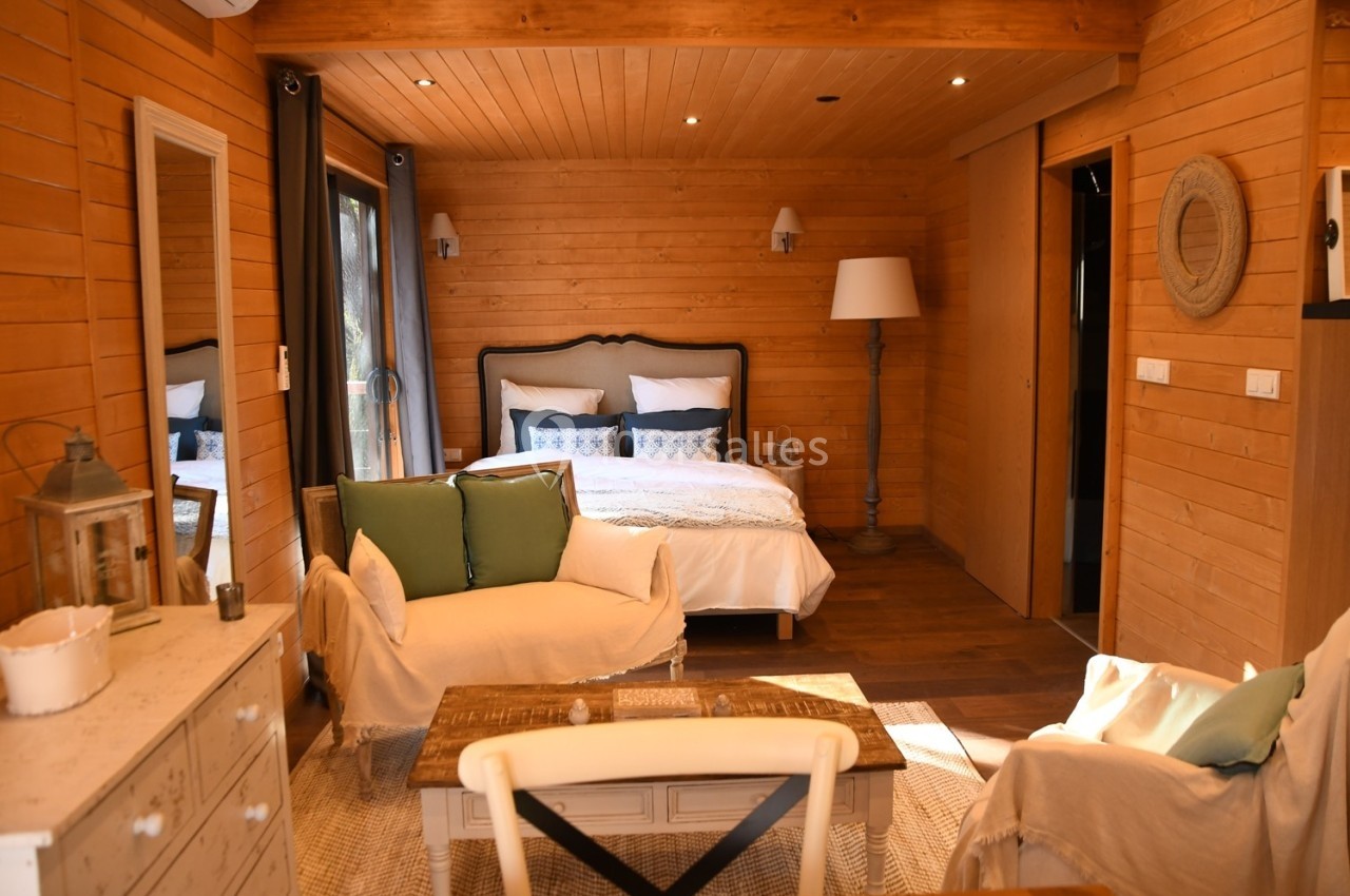 Chambre en bois avec lit double, fauteuils, table basse et lumière naturelle entrant par une baie vitrée.
