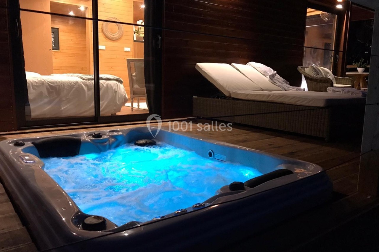 Bain à remous éclairé en bleu sur une terrasse en bois, avec vue sur une chambre et des transats en arrière-plan.