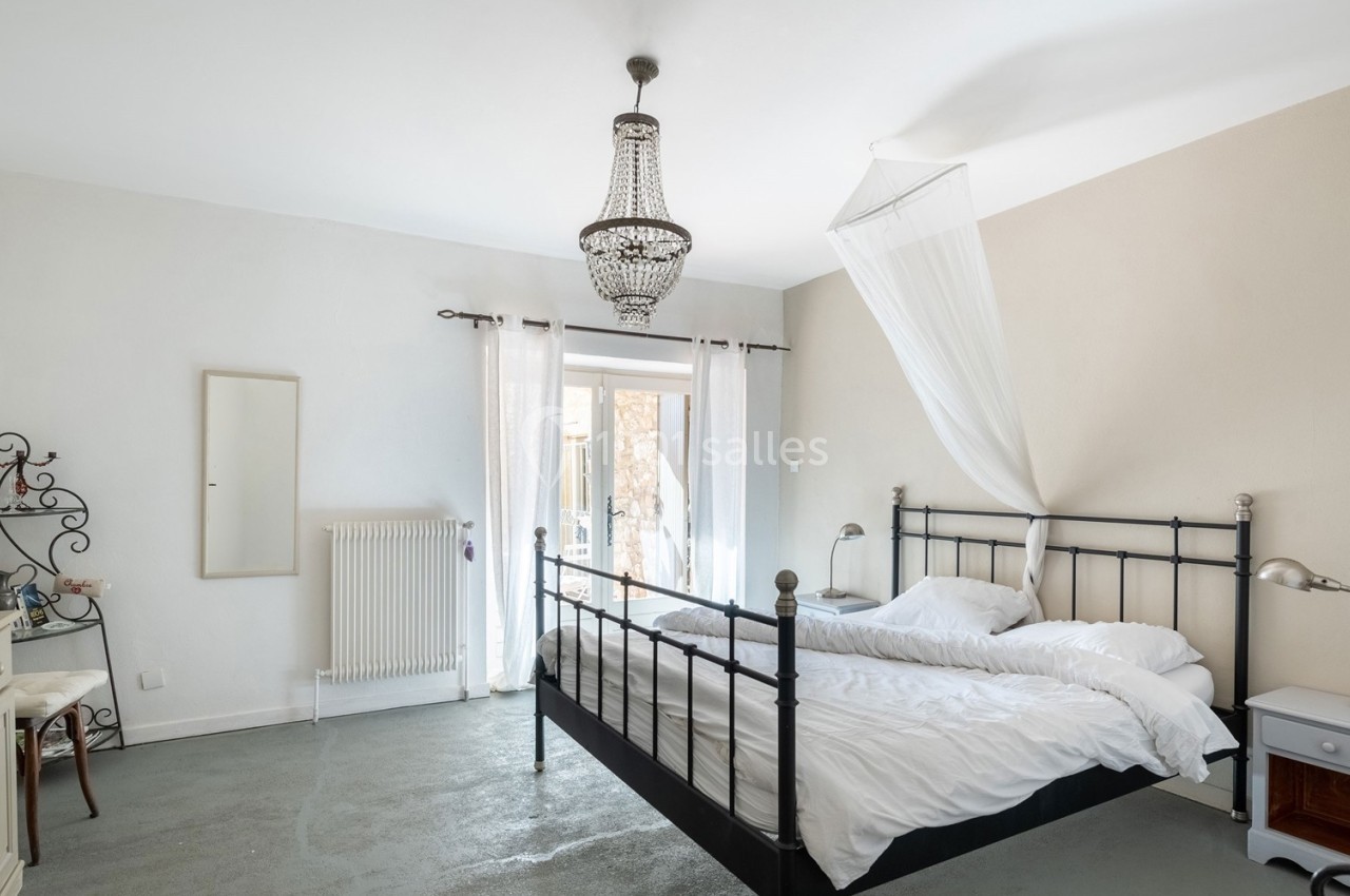 Chambre lumineuse avec lit double en métal noir, moustiquaire, lustre et accès à une porte-fenêtre donnant sur l'extérieur.