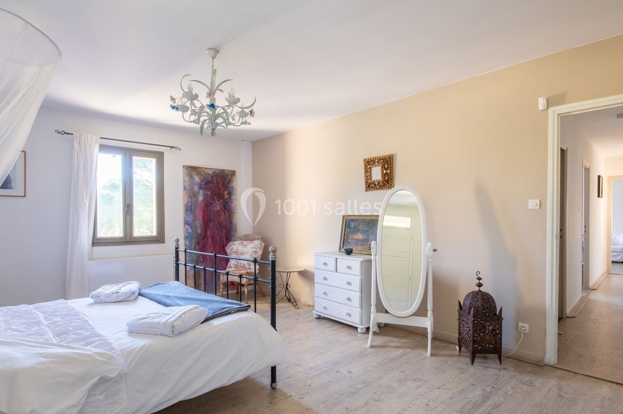 Chambre lumineuse avec lit double, commode blanche, miroir sur pied et vue sur un couloir à droite.