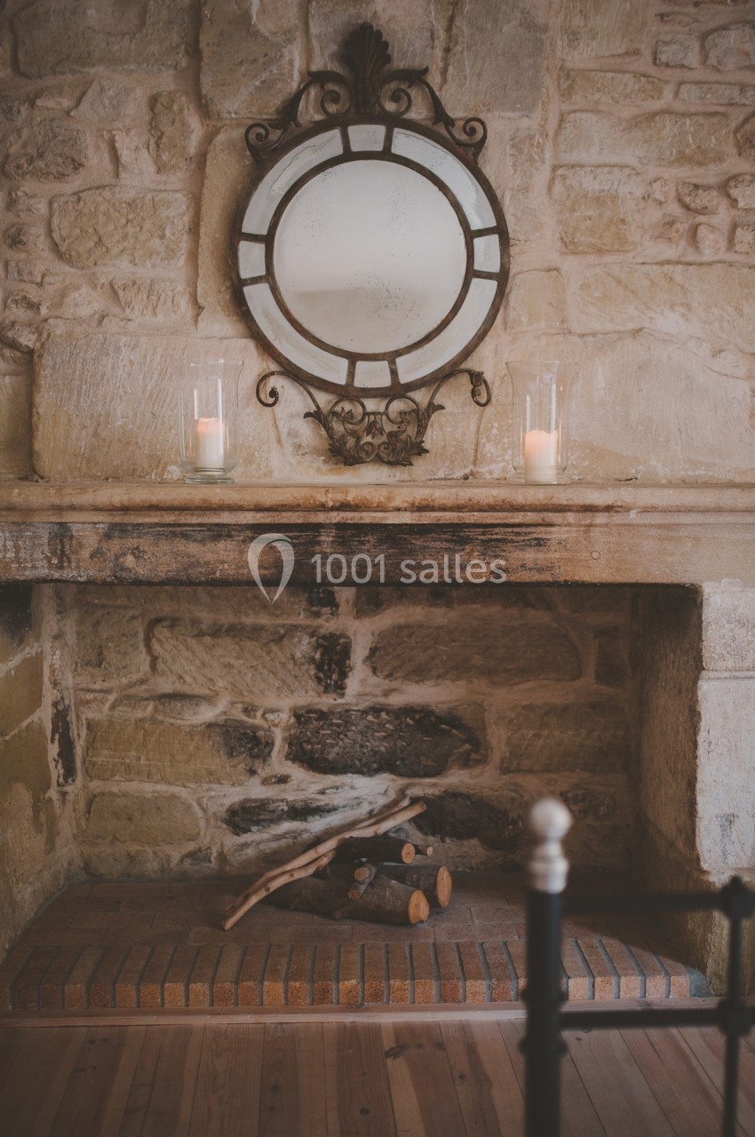 Cheminée en pierre avec miroir rond encadré en métal, deux bougies allumées et quelques bûches posées en dessous.
