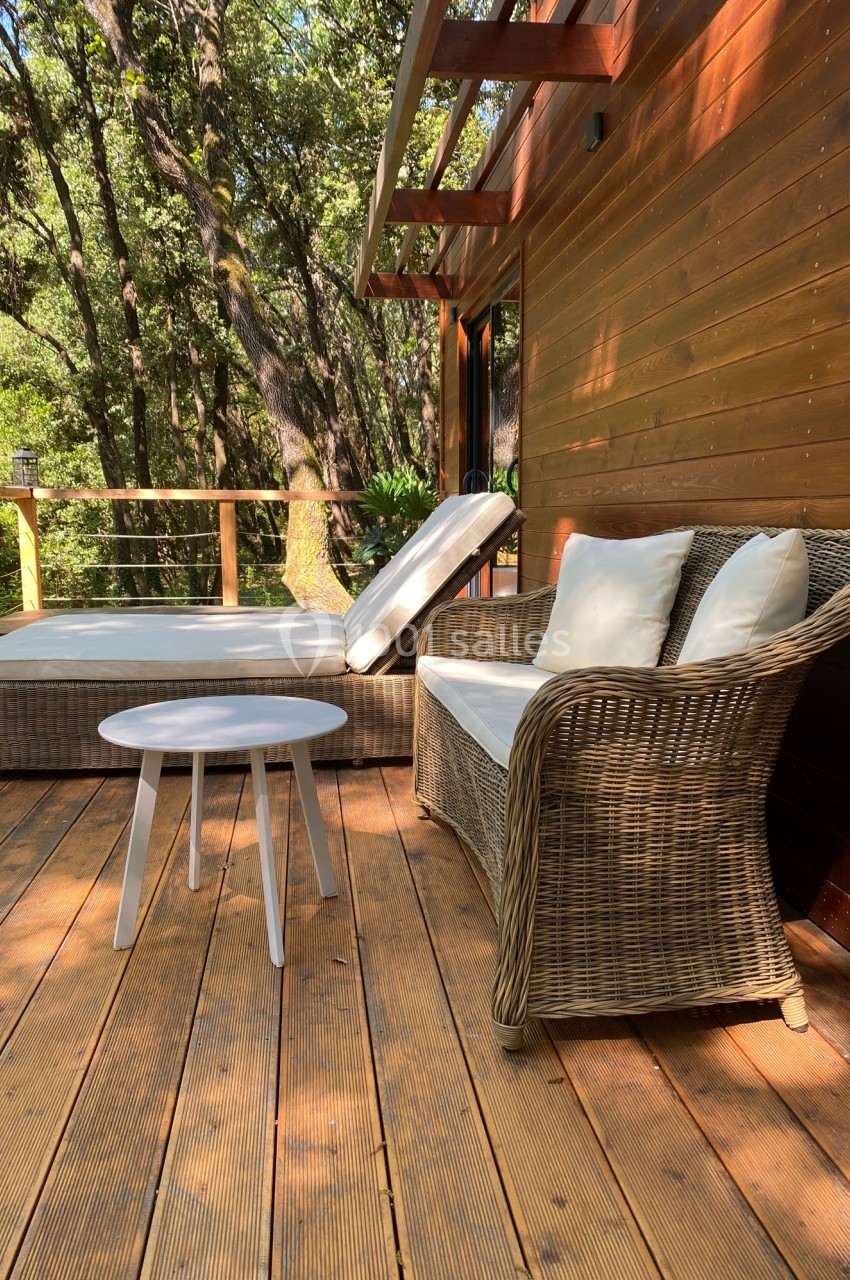 Terrasse en bois avec fauteuil en rotin, table basse blanche et transat, entourée d'arbres et de verdure.