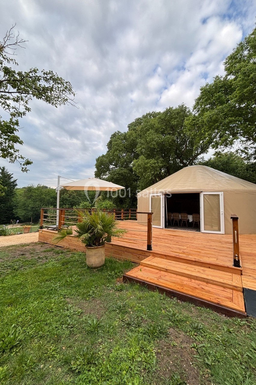 Yourte beige avec terrasse en bois, parasol blanc et végétation environnante sous un ciel partiellement nuageux.