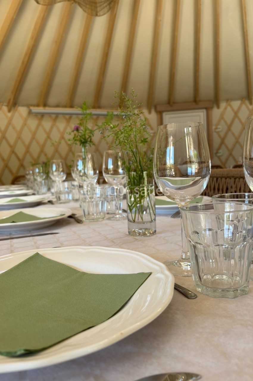 Table dressée avec des assiettes blanches, serviettes vertes, verres et décorations florales dans un cadre en bois clair.