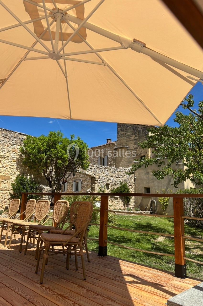 Terrasse en bois avec chaises en rotin sous un grand parasol beige, vue sur un jardin et des bâtiments en pierre.