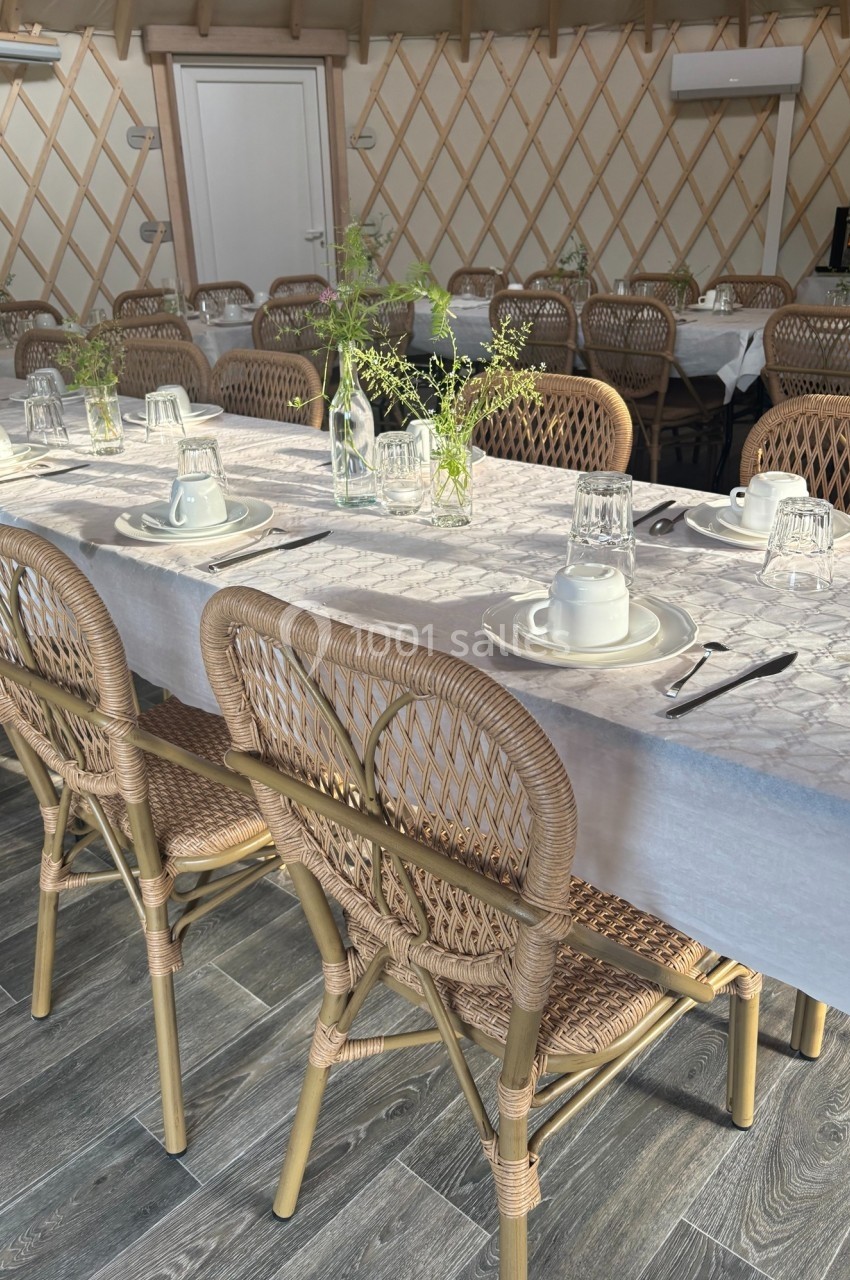 Salle aménagée avec des tables dressées, nappes blanches, chaises en rotin et décoration florale simple.