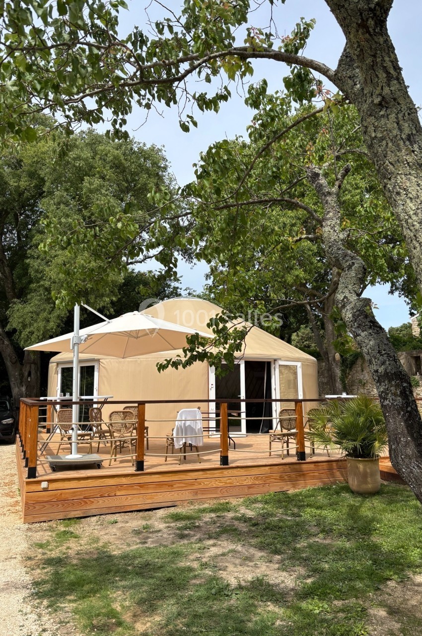 Tente de style yourte avec terrasse en bois, entourée d'arbres et de végétation, par une journée ensoleillée.