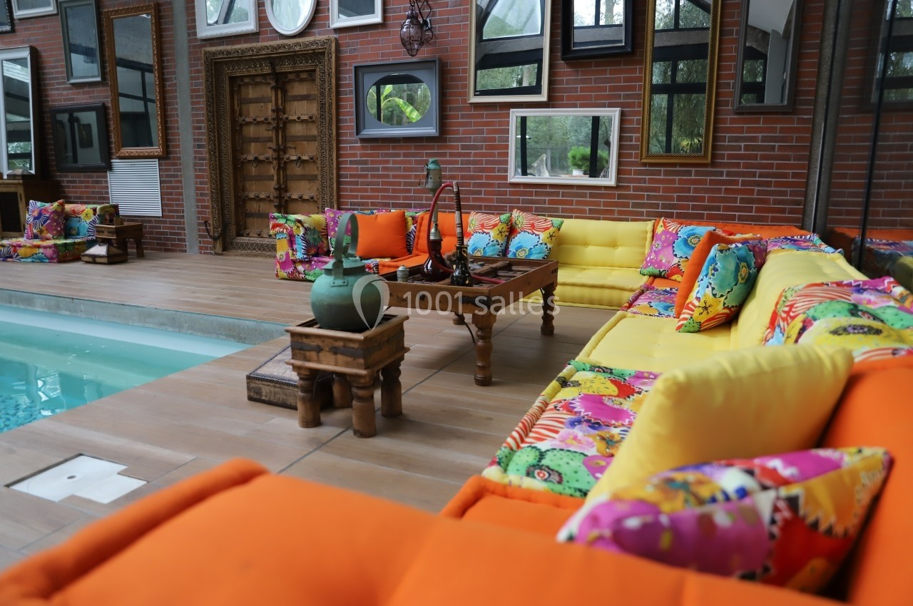 Salon extérieur coloré avec canapés orange et jaunes, coussins à motifs vifs, table basse en bois et piscine adjacente.