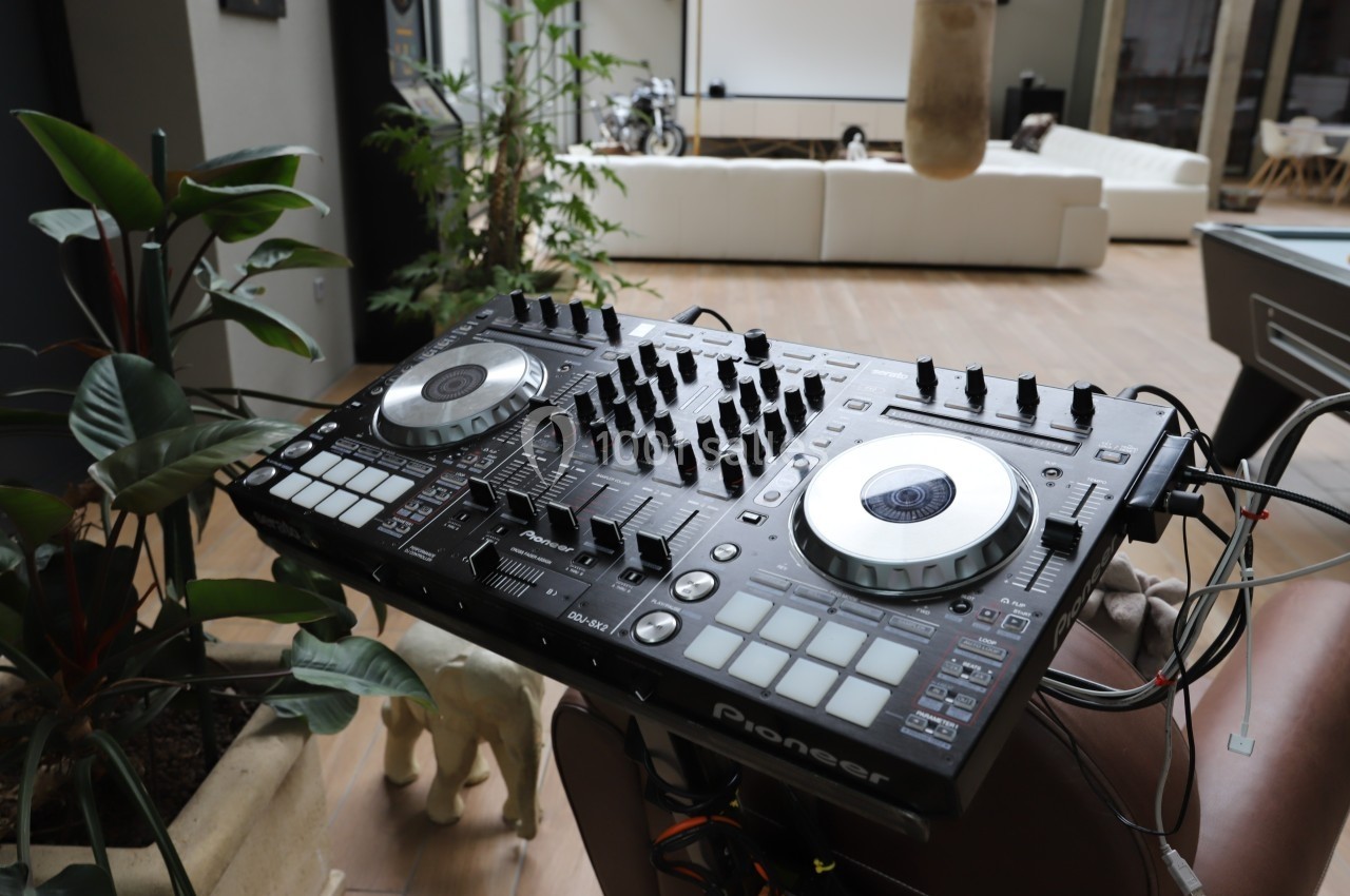 Table de mixage DJ avec platines et commandes, installée dans un espace intérieur moderne avec des plantes et un canapé.