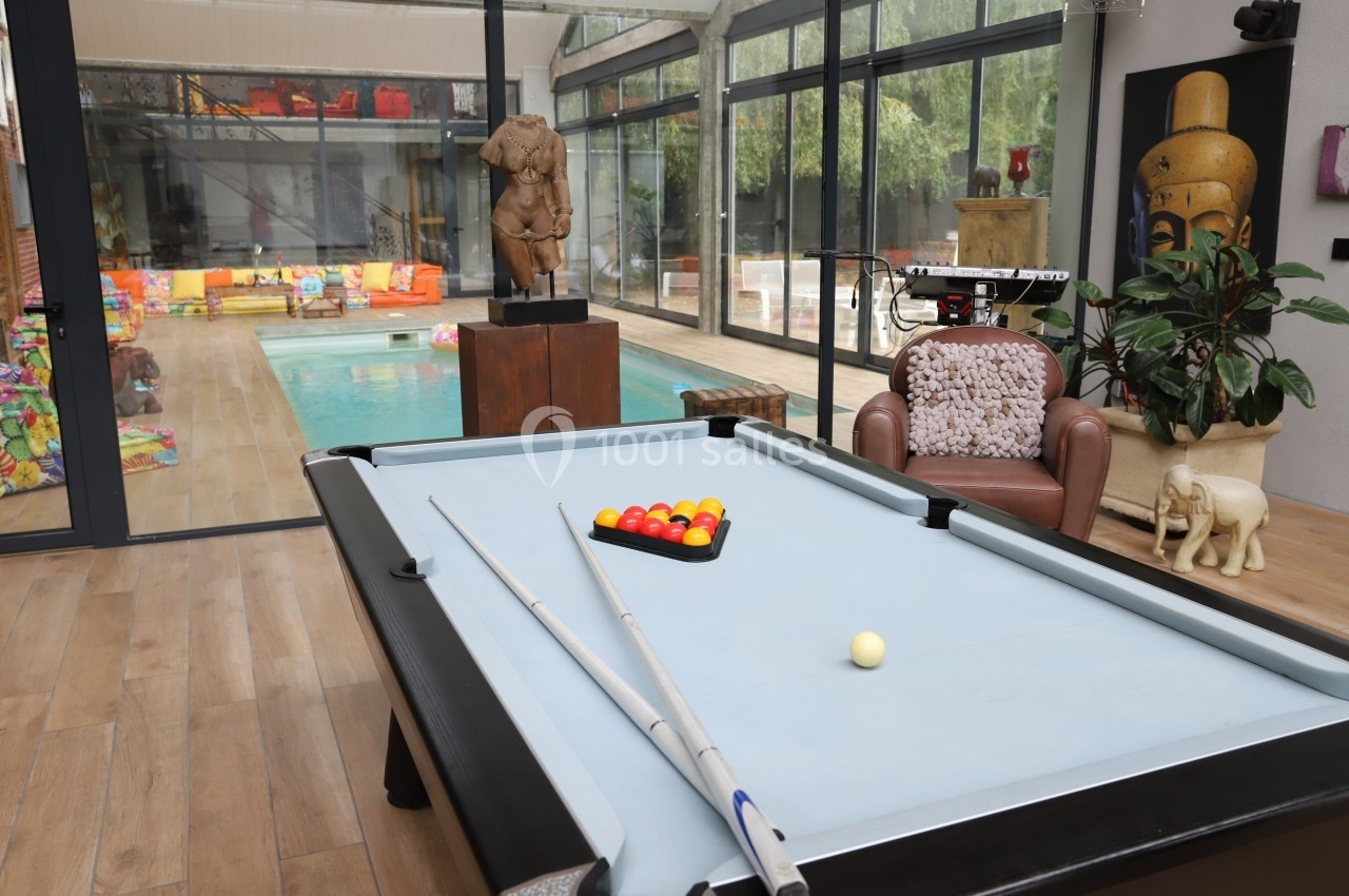 Table de billard avec accessoires au premier plan, piscine intérieure et décoration variée en arrière-plan.