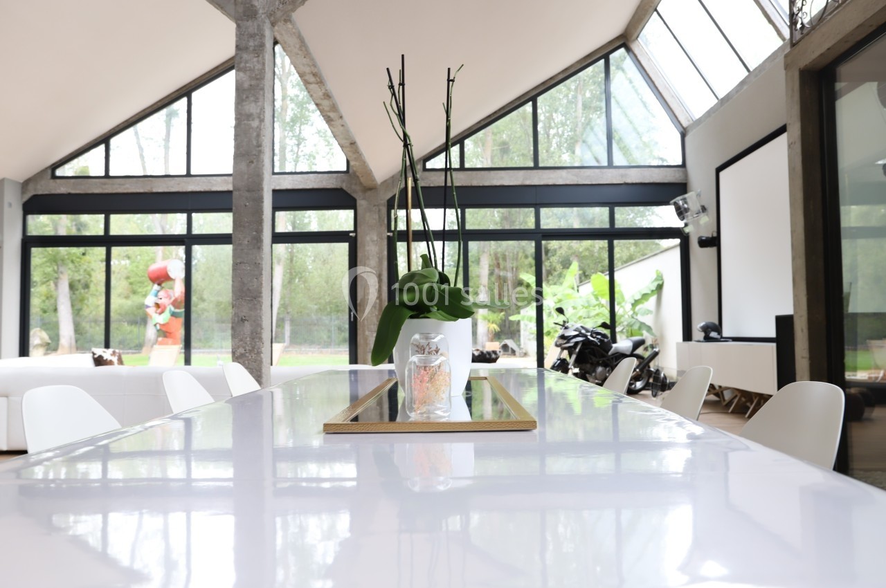 Salle lumineuse avec grande table blanche, vase contenant une plante au centre, baies vitrées et vue sur jardin.