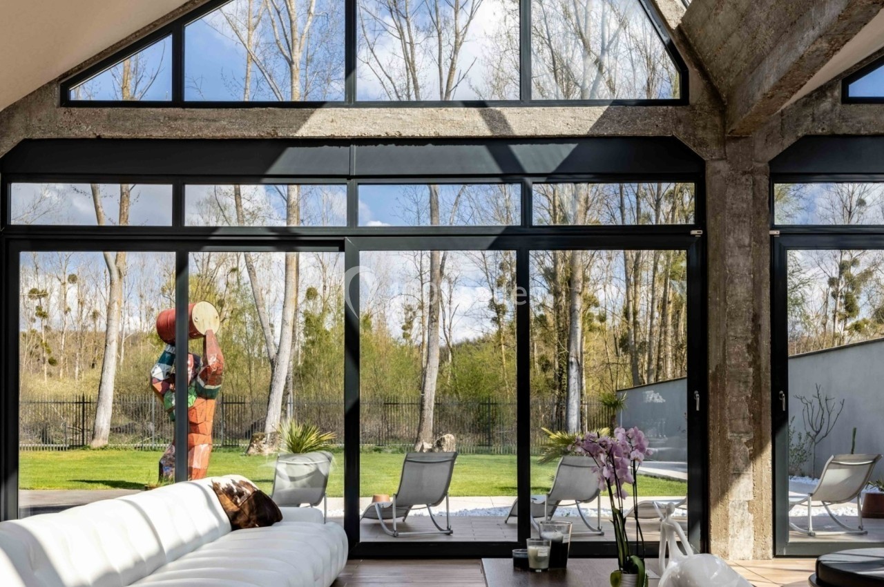 Intérieur lumineux avec grandes baies vitrées donnant sur un jardin arboré, fauteuils extérieurs et sculpture colorée.