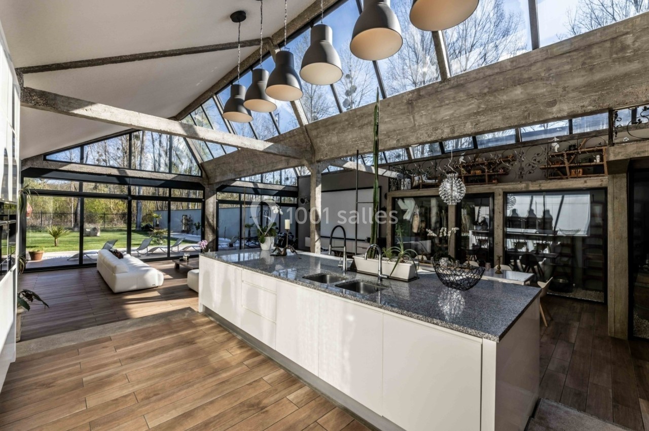 Cuisine moderne avec îlot central, grandes baies vitrées, plafond en pente et vue sur un jardin arboré.