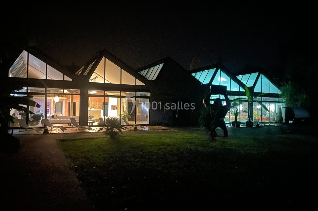 Vue nocturne d'une maison moderne avec de grandes baies vitrées illuminées donnant sur un jardin sombre.
