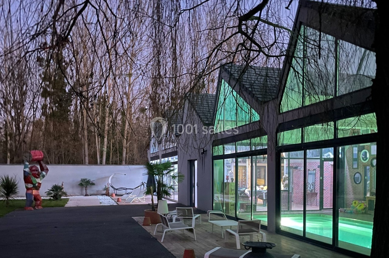 Façade vitrée d'un bâtiment avec piscine intérieure éclairée, entourée de chaises longues et d'arbres en arrière-plan.