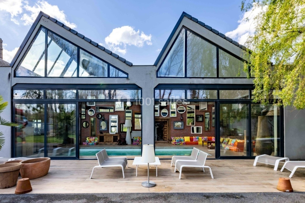 Façade vitrée d'une maison moderne avec terrasse en bois, mobilier extérieur blanc et vue sur un intérieur coloré.