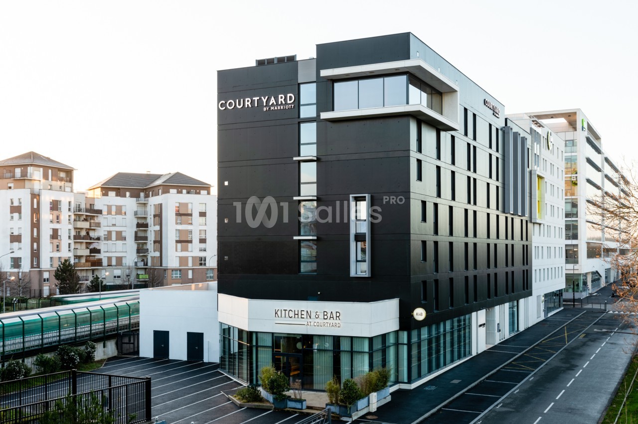 Façade d'un hôtel moderne de sept étages en panneaux sombres, avec un rez-de-chaussée vitré abritant un restaurant, sous un ciel clair, à côté d'immeubles d'habitation. Wait, the output must be one sentence, ≤125 characters. Let me count and shorten.