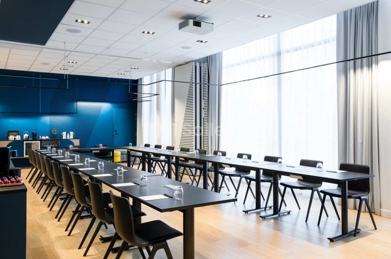 Salle de réunion en configuration en U avec tables noires, chaises noires et verres d'eau, murs bleus, grandes baies vitrées et vidéoprojecteur au plafond.