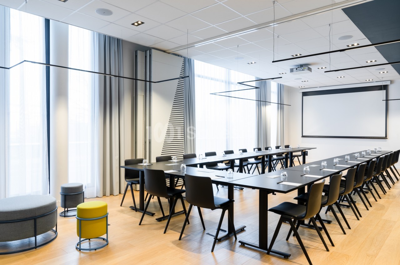 Salle de réunion en configuration en U avec tables noires et chaises noires, écran de projection au fond, grandes baies vitrées et parquet clair.