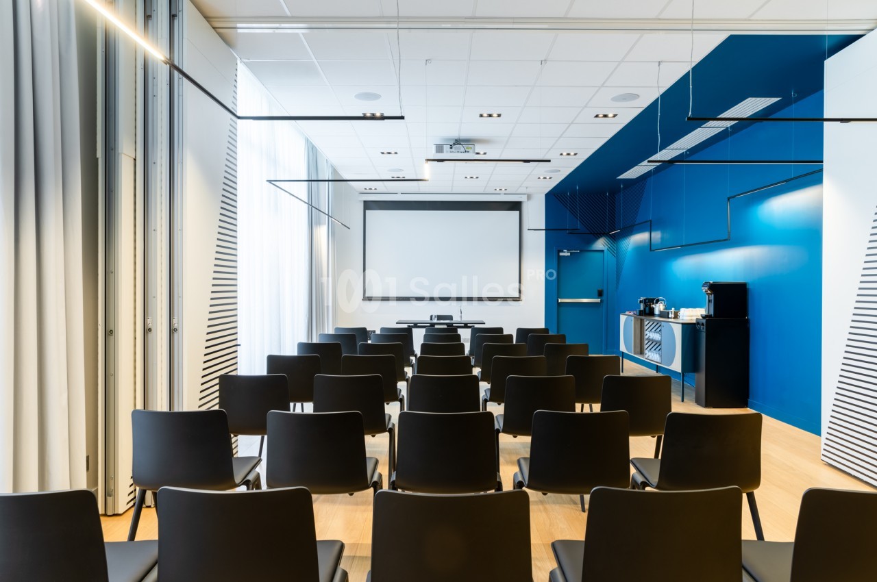 Salle de réunion en configuration théâtre, rangées de chaises noires face à un écran de projection, avec un mur bleu et un buffet à droite.