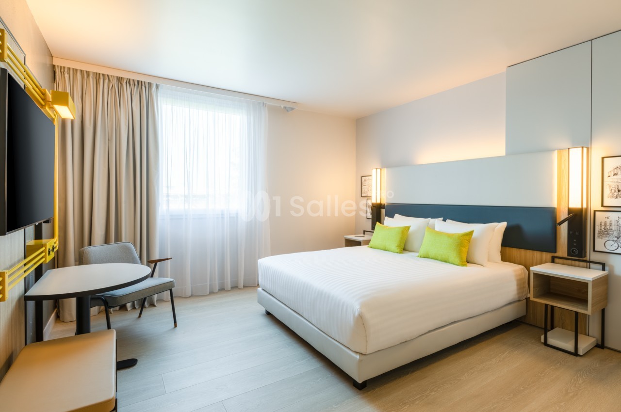 Chambre d'hôtel avec grand lit blanc aux coussins verts, bureau rond avec deux fauteuils, téléviseur mural et grande fenêtre aux voilages clairs.