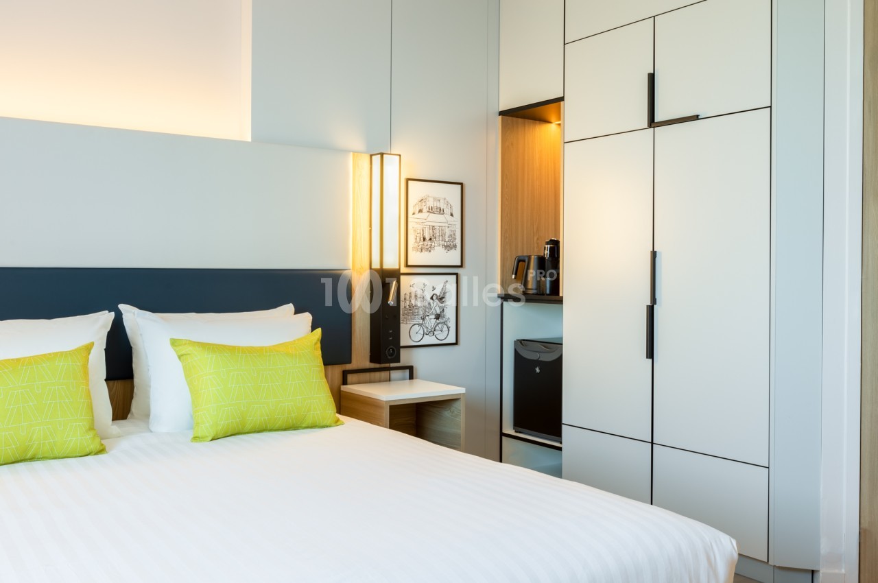 Chambre d'hôtel avec un grand lit aux draps blancs, coussins verts, tête de lit sombre, lampe murale et armoire encastrée blanche avec plateau de café. Let me double-check length: about 165 characters, too long. Need ≤125. Chambre d'hôtel au lit blanc
