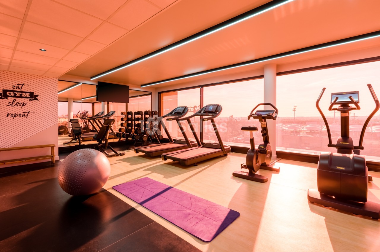 Salle de sport avec deux tapis de course, un vélo d'appartement et un vélo elliptique face à de grandes baies vitrées, un tapis de yoga violet au sol.