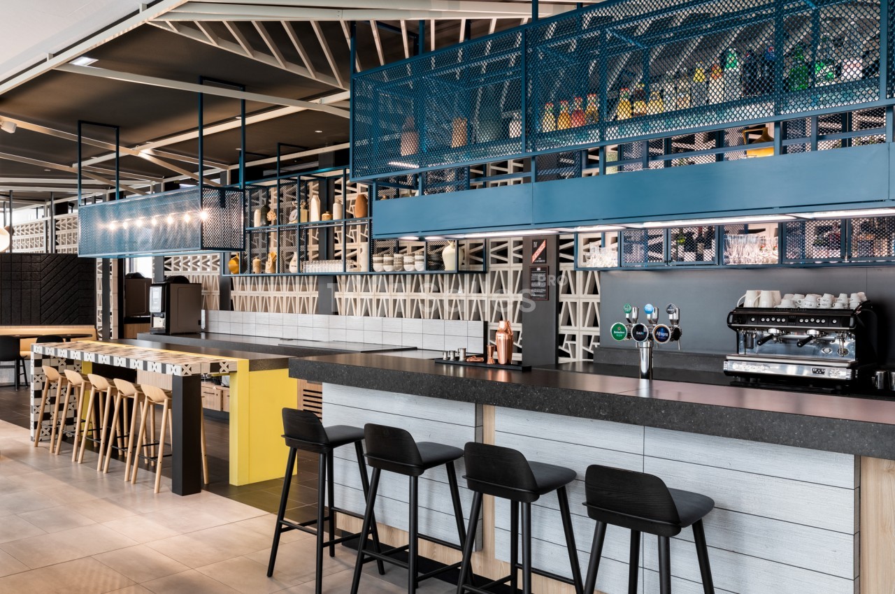 Bar d'hôtel moderne avec comptoir gris foncé, tabourets noirs, machine à espresso, tireuses et étagère métallique bleue garnie de bouteilles.